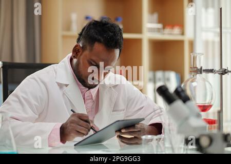 Serio clinico o ricercatore afroamericano che utilizza tablet dal posto di lavoro in laboratorio, mentre la ricerca di informazioni online su nuovi virus Foto Stock