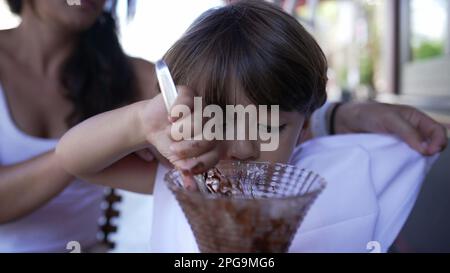 Madre che mette il tovagliolo intorno al collo del bambino per proteggere dalle macchie mentre mangia il dessert del gelato del cioccolato con il cucchiaio Foto Stock