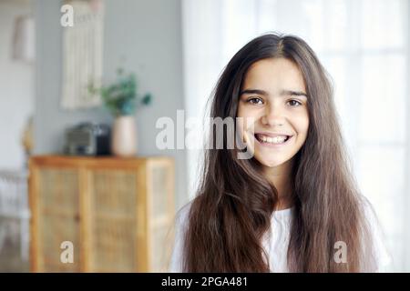 Seria bella preseen 12s ragazza posa in accogliente soggiorno, avendo lungo brunette capelli sorriso guardando la macchina fotografica. Generazione Alpha persona ritratto Foto Stock