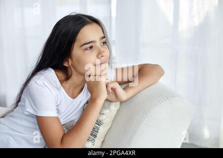 Bella ragazza 12s presen pensivo sedersi sulla poltrona in accogliente salotto alla moda guardando in lontananza e pensiero. Generazione Z ritratto persona, d Foto Stock