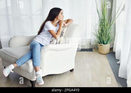 Bella ragazza 12s presen pensivo sedersi sulla poltrona in accogliente salotto alla moda guardando in lontananza e pensiero. Generazione Z ritratto persona, d Foto Stock