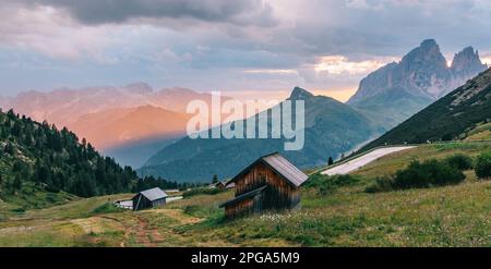 Tranquillo paesaggio alpino delle Alpi italiane, calda luce arancione dorata del sole del mattino illumina le pittoresche case in legno e prati verdeggianti o Foto Stock