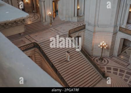 Una coppia ha visto in piedi mano presso la grande scalinata del Municipio di San Francisco. La sposa sta beaming con felicità, e lo sposo la sta guardando con amore e ammirazione. Gli elementi storici dell'edificio forniscono un ambiente bello e memorabile per il giorno del matrimonio della coppia. Dopo due anni di lavori di ristrutturazione, l'iconico municipio di San Francisco ha finalmente riaperto le sue porte al pubblico dal giugno 2021. Il progetto da $550 milioni di dollari mirava a ripristinare gli elementi storici dell'edificio e a modernizzarlo per soddisfare le esigenze del 21st° secolo. Il progetto prevedeva il restauro della Cit Foto Stock