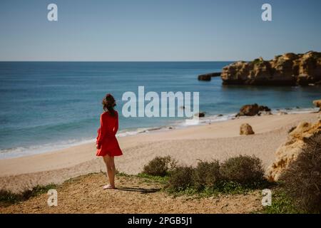 Donna attraente in vestito rosso sulla cima delle scogliere a Sao Rafael Beach, Costa dell'Algarve, Portogallo Foto Stock