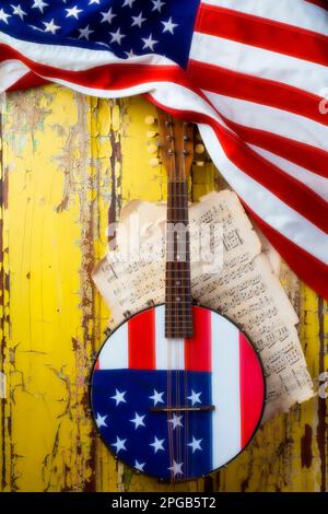 American Banjo con American Flag, appeso su una rustica vernice alle intemperie che sbuccia la porta gialla con musica spartiti consumata. anni '20 banjo arte popolare ancora vita. Foto Stock