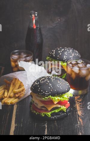 Set di hamburger neri con polpettine di carne, formaggio, pomodori, maionese, patatine fritte in una tazza di carta e bicchiere di cola fredda soda con ghiaccio. Legno scuro Foto Stock