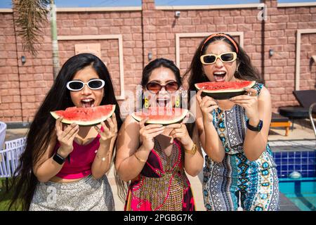 Tre belle giovani ragazze indiane che si divertono insieme e mangiare anguria nel caldo giorno d'estate, amici che indossano occhiali da sole che tengono una fetta di f tropicale Foto Stock