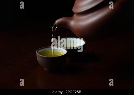 Due tazze bianche in ceramica con tè verde fumoso che vengono versate in loro da una teiera su uno sfondo scuro Foto Stock