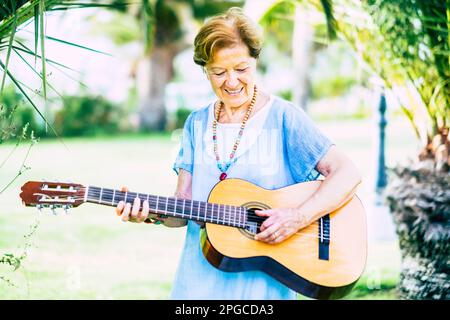 Donna anziana con problemi di salute come demenza senile o alzheimer suonando da sola una chitarra al parco in assistenza casa matura ospizio. reti felice Foto Stock