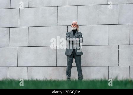 Uomo d'affari senior in abiti formali, con capelli grigi e barba è all'aperto si trova contro il muro Foto Stock