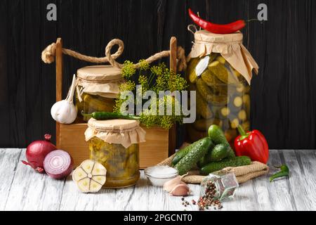 Cetrioli sottaceto in vaso, una scatola di legno con conservazione su fondo di legno scuro. Peperoni, cipolle e cetriolini freschi per la conservazione. Cetriolo Foto Stock