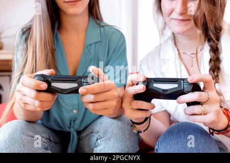 Le amiche femminili sono irriconoscibili e si rilassano su un comodo divano di colore rosso con controller per videogiochi in mano e si divertono con la dur del tempo libero Foto Stock
