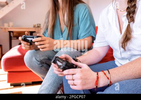 Le amiche femminili sono irriconoscibili e si rilassano su un comodo divano di colore rosso con controller per videogiochi in mano e si divertono con la dur del tempo libero Foto Stock