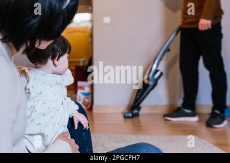 Madre irriconoscibile seduta con bambina e padre piano di pulizia con aspirapolvere in background a casa Foto Stock