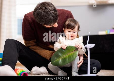 Allegro padre in abiti casual seduto sul pavimento con cute figlia giocare con foglia verde, mentre trascorrere il tempo insieme a casa Foto Stock