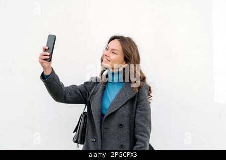 Allegra giovane donna in cappotto con borsa che si autoritratto su smartphone e sorridente su sfondo bianco Foto Stock