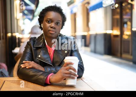 Giovane ragazza afroamericana in giacca di pelle bere caffè e guardare la macchina fotografica mentre si siede al tavolo di legno in Street cafe Foto Stock