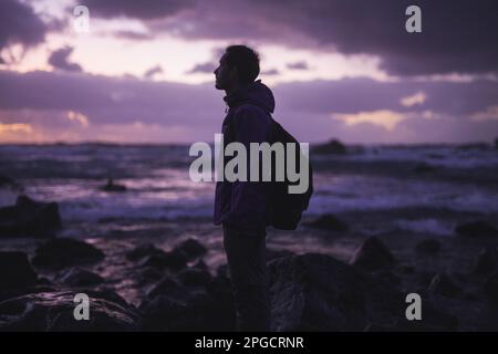 Vista laterale del giovane viaggiatore maschio con zaino in piedi sulla costa rocciosa vicino mare ondeggiante contro cielo nuvoloso tramonto nelle Azzorre, Portogallo Foto Stock