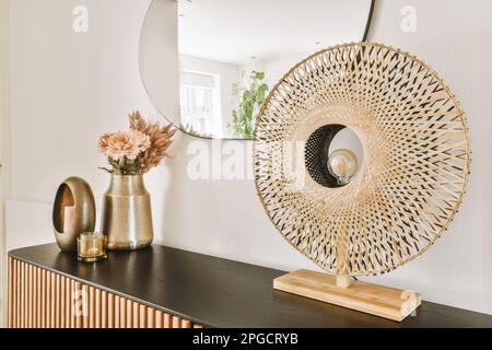 Lampada da tavolo in legno e bouquet di fiori freschi in vaso posto su tavolo in legno con specchio sopra contro parete bianca Foto Stock