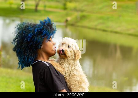 Goiania, Goias, Brasile – 20 marzo 2023: Una giovane donna nera con capelli blu tinti, con il cane in grembo, in un parco ben boscoso. Foto Stock