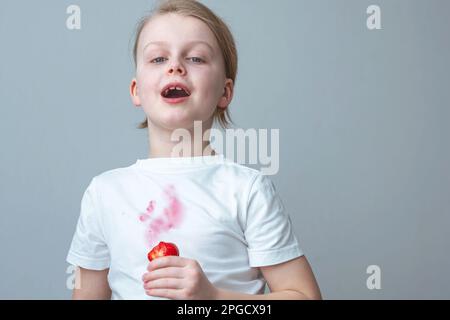 Ritratto di un ragazzo allegro con fragole fresche in mano su sfondo grigio. Una macchia rosa sporca sulla parte anteriore di una t-shirt bianca. Foto Stock