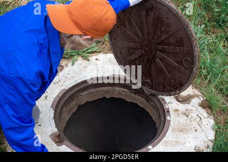 Un lavoratore apre la copertura del tombino e guarda verso il basso nel pozzo. Lavori idraulici per controllare e pulire i pozzi settici. Foto Stock