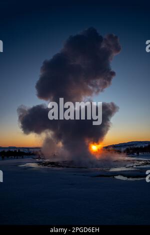 Il grande geyser Strokkur dell'Islanda in piena eruzione con nebbia e fumo retroilluminati e la sera arancione o il sole vicino al tramonto che tocca l'orizzonte dietro Foto Stock