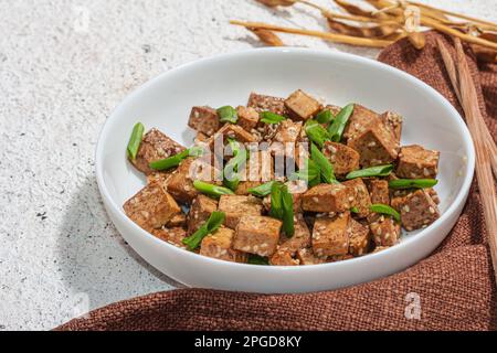 Tofu teriyaki fritto con scalioni e semi di sesamo. Pasto vegano sano senza carne ricco di proteine e calcio. Luce dura alla moda, ombra scura, intonaco Foto Stock