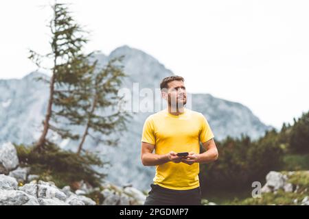 Vita in su l'uomo caucasico in una camicia gialla che guarda in su nel cielo mentre tiene il telefono nelle sue mani, controllando le previsioni del tempo in su nelle montagne Foto Stock