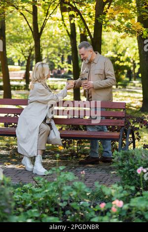 Uomo sorridente che dà la tazza di carta con il caffè alla moglie bionda sulla panca in parco, immagine di scorta Foto Stock