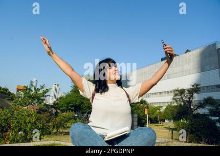 giovane studentessa latina venezuelana, seduta a braccia aperte all'aperto guardando il suo telefono cellulare molto felice per i suoi successi, con l'università dietro di lui Foto Stock