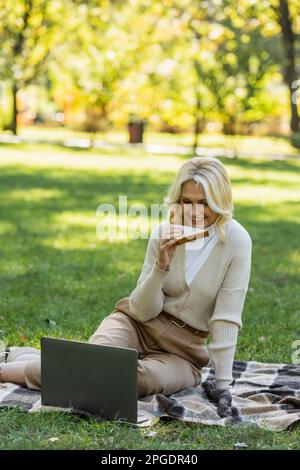 donna felice di mezza età con i capelli biondi mangiare sandwich e guardare film sul computer portatile durante il pic-nic nel parco, immagine stock Foto Stock