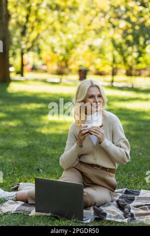 felice donna di mezza età che mangia il panino del randello e che siede sulla coperta vicino al laptop durante il picnic nel parco, immagine di riserva Foto Stock