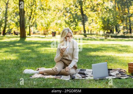 allegra donna di mezza età con capelli biondi mangiare sandwich e guardare film sul computer portatile durante il picnic nel parco, immagine stock Foto Stock