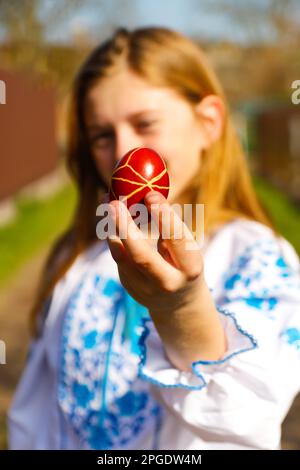 Defocus ragazza ucraina in vyshyvanka tenendo un uovo rosso colorato su sfondo naturale. Pasqua, Ucraina. Uova dipinte artigianalmente. Pysanka o krashanka. Sfocatura Foto Stock