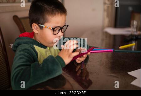 Primo piano ritratto di un ragazzo che guarda nel telefono seduto al tavolo, giocando al telefono invece di disegnare con matite Foto Stock