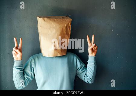 Felice uomo positivo con un sacchetto di carta mostra segno di vittoria su sfondo grigio. Concetto di emozione della gente Foto Stock