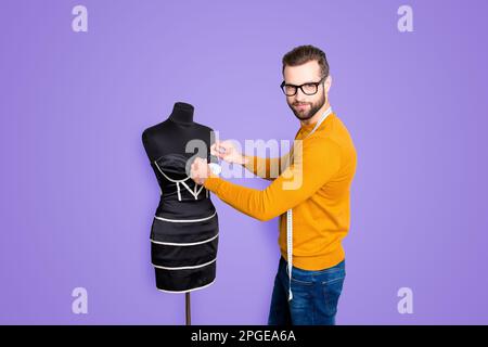 Ritratto di bella comò attraente in maglione e jeans con centimetro sul collo prendendo misure da abito su manichino con aghi guardare Foto Stock