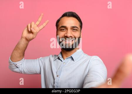 Gioioso giovane uomo indiano sta utilizzando lo smartphone per le videochiamate, sorridente ragazzo comunicare in internet, registrazione vlog, godersi la conversazione online isolato sul rosa, prende selfie, mostrando v-segno nella fotocamera Foto Stock