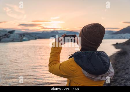 Turista femminile in giacca gialla scattando foto con smartphone nel ghiacciaio della laguna di Jokulsarlon in Islanda Foto Stock