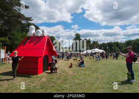 Persone che prendono selfie con i loro cani in piedi di fronte a un enorme allevamento rosso con Snoopy sdraiato in cima a Goodwoof, The Kennels, Goodwood, Sussex, UK Foto Stock