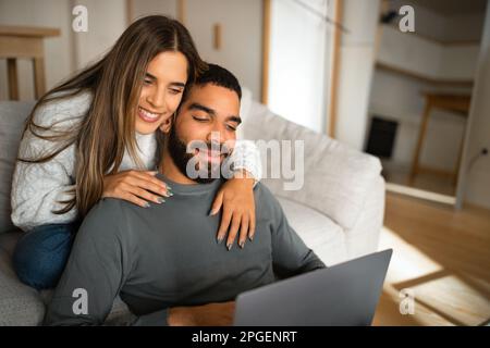 Allegra coppia millenaria multietnica sedersi sul divano, abbracciando, guardando il computer portatile, hanno videochiamata Foto Stock