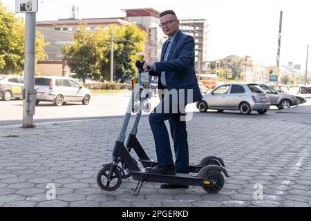 Giovane uomo d'affari in tuta in sella a uno scooter elettrico in una riunione d'affari. Concetto di trasporto ecologico, noleggio di uno scooter elettronico sul ci Foto Stock