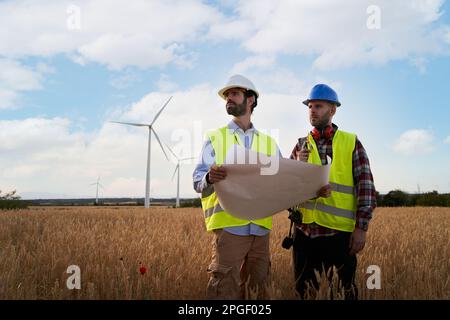 Due ingegneri in cappellini duri lavorano in aereo in campo agricolo con turbine eoliche. All'aperto. Foto Stock