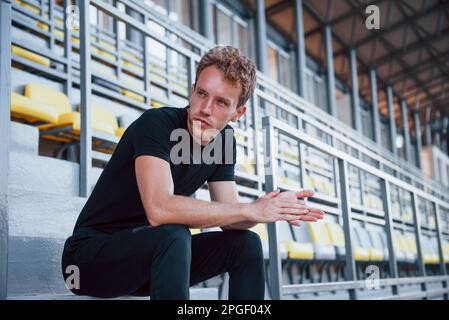 Si siede sulle scale. Giovane sportivo in camicia nera e pantaloni all'aperto di giorno Foto Stock