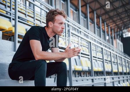 Si siede sulle scale. Giovane sportivo in camicia nera e pantaloni all'aperto di giorno Foto Stock