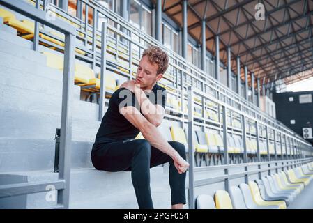 Si siede sulle scale. Giovane sportivo in camicia nera e pantaloni all'aperto di giorno Foto Stock