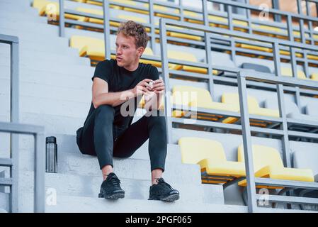 Si siede sulle scale. Giovane sportivo in camicia nera e pantaloni all'aperto di giorno Foto Stock