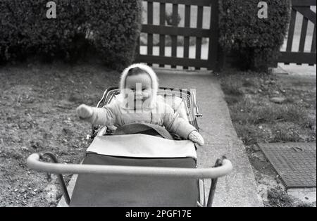 1960s, storico, un giovane bambino bonny seduto in un pram parcheggiato su un percorso concreto all'esterno ad un'entrata ad una casa, Inghilterra, Regno Unito. Foto Stock