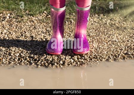 Bambino che indossa nuovi stivali di gomma da pioggia rosa brillante in piedi vicino a pozza fangosa, senza volto. Prescolare in wellies. Calzature per il tempo piovoso Foto Stock